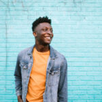 A smiling African American man stands in front of a blue wall wearing nice casual clothing.  Positive lifestyle portrait.  Shot in Portland, OR, USA.